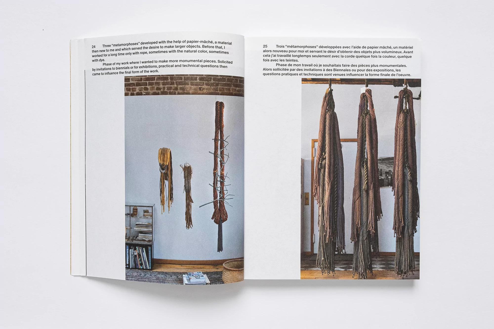 Photograph inside an open book featuring paper mâché art installations against white walls, including fiber sculptures and shelves of books.