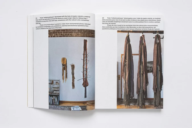 Photograph inside an open book featuring paper mâché art installations against white walls, including fiber sculptures and shelves of books.