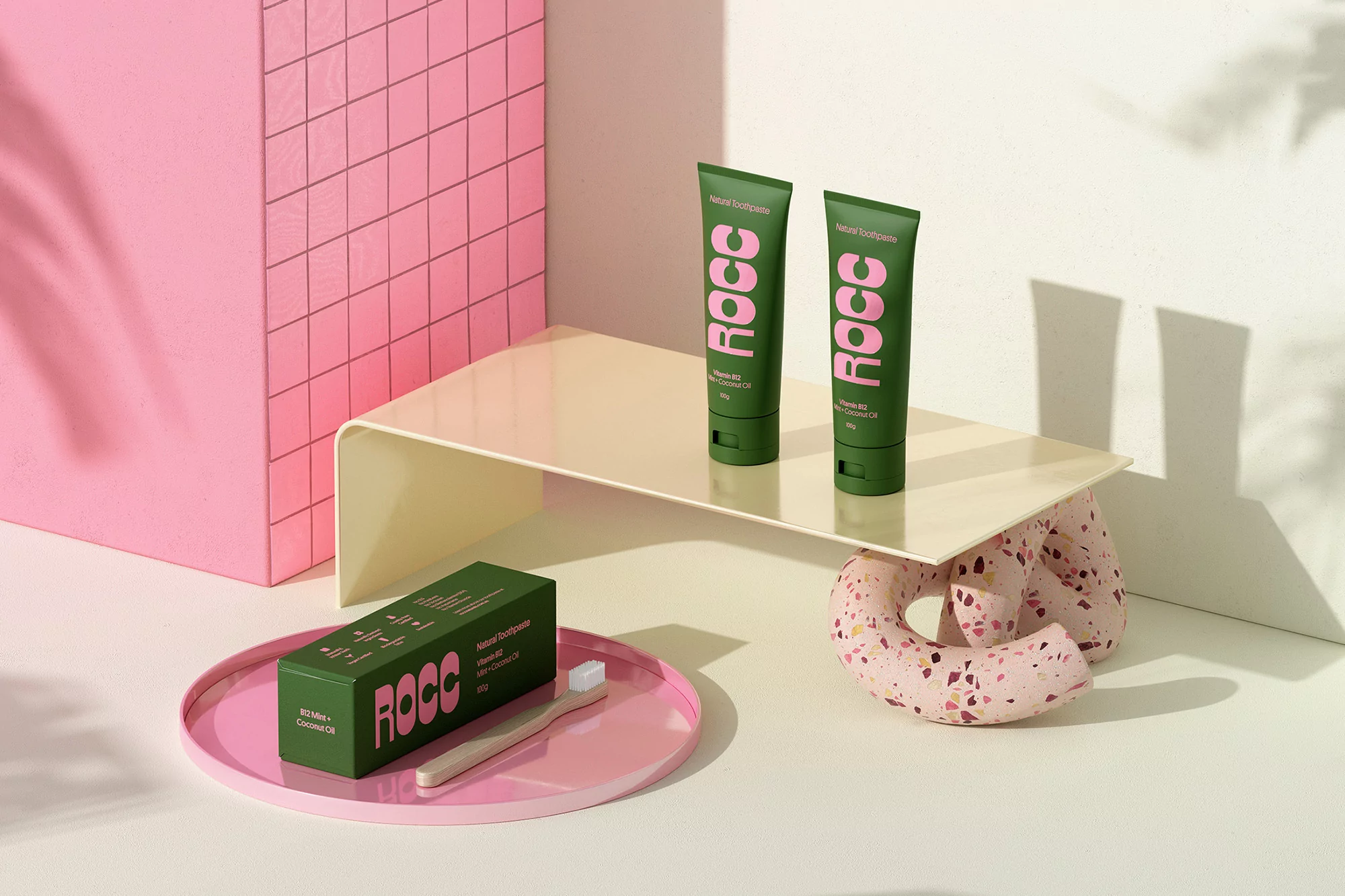 Studio shot of two green tubes and a box of toothpaste, next to a toothbrush on a pink plate, creating a clean beauty aesthetic.
