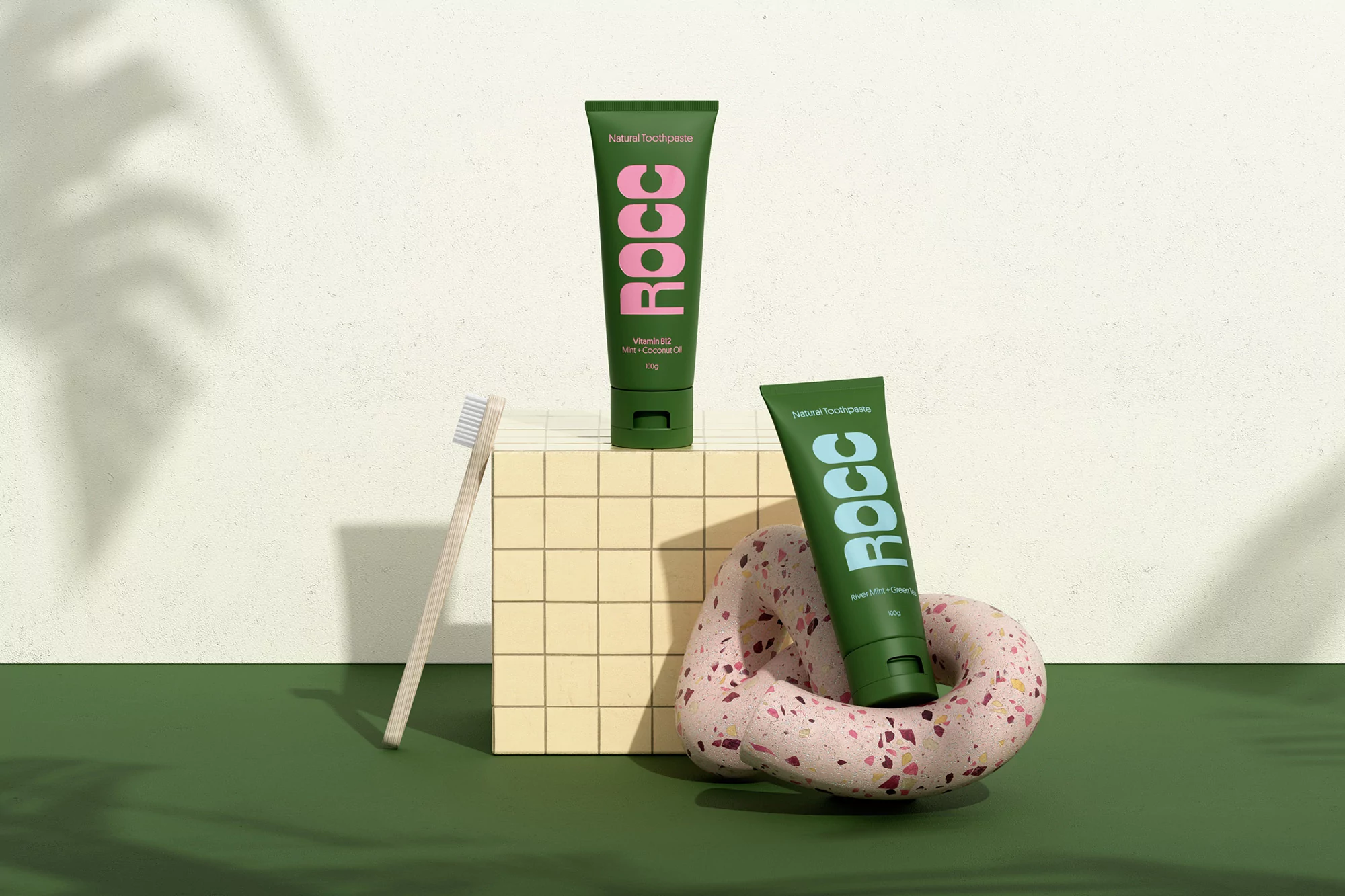 Product shot of green ROCC natural toothpaste tubes with pink and light blue logos, toothbrush, and decorative stone.