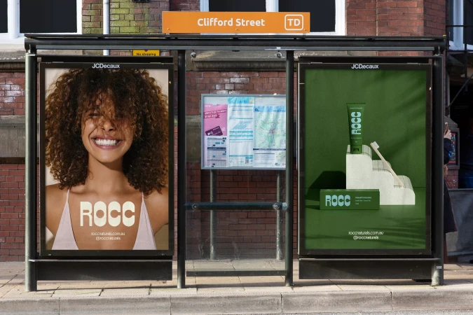 Outdoor bus stop advertisement featuring a smiling woman with curly hair alongside an ad for ROCC brand toothpaste and toothbrush.