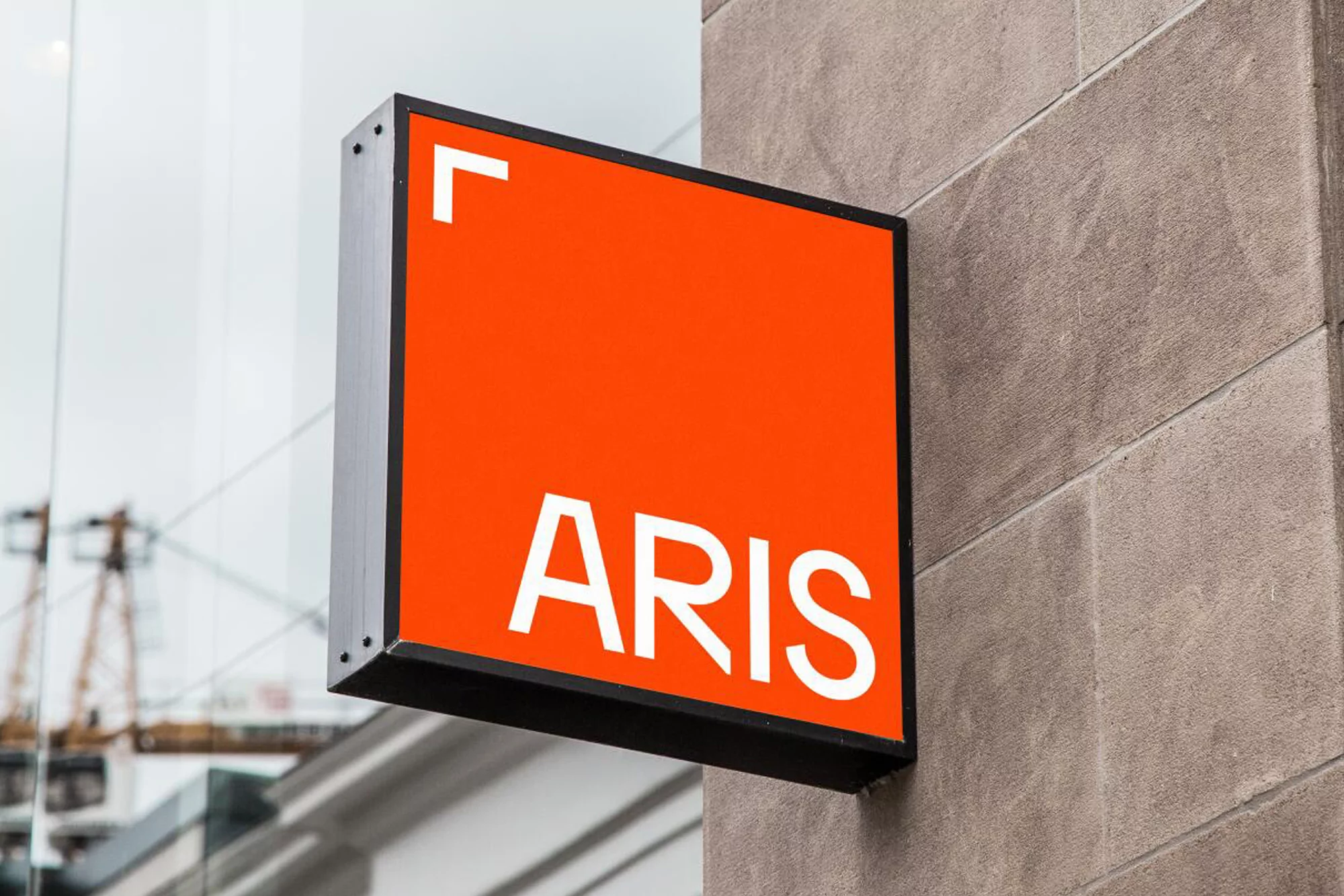 Close-up of a square orange sign with the white text 'ARIS' attached to the exterior of a building with a construction site reflection.