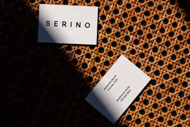 Two white business cards resting on a textured woven rattan surface with diagonal shadows cast across them.