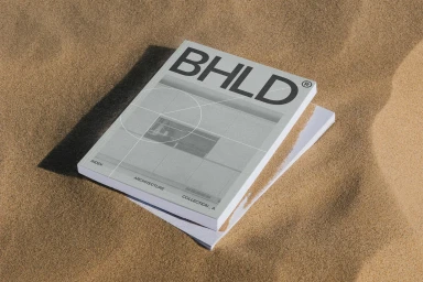 A book titled BHLD resting on rippled sand dunes under direct sunlight casting a sharp shadow.