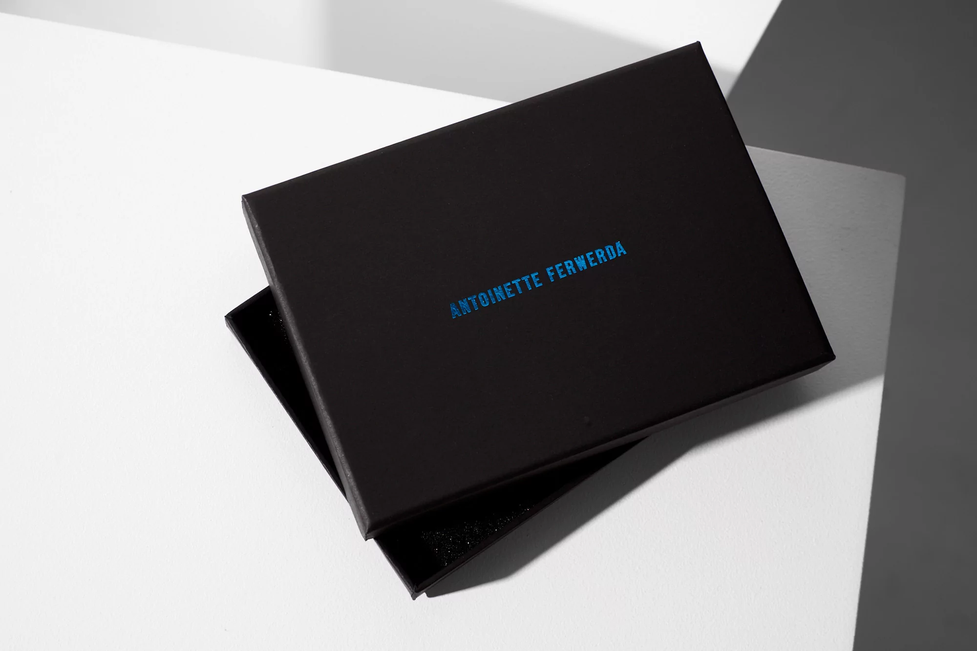Elevated view of a black rectangular box with the text 'Antoinette Ferwerda' in blue font, placed on a clean white surface.