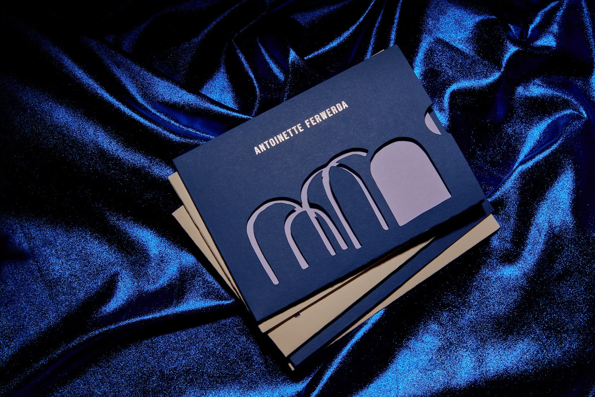 Stationery set with a dark blue business card featuring a unique geometric cut-out design, resting on a shimmering blue satin fabric backdrop.