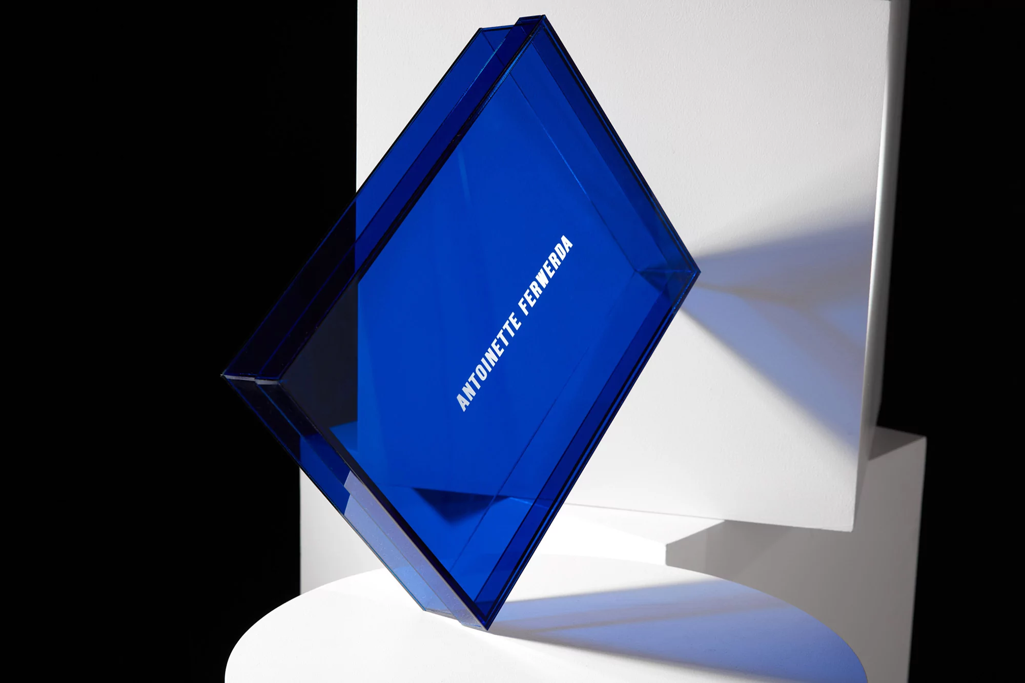 Studio shot of a translucent blue prism with the text 'Antoinette Ferwerda' on a white geometric platform against a black background.