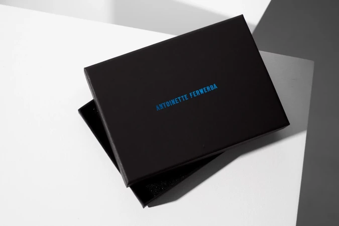 Black gift box with blue branded text resting on angular white and grey studio surfaces.