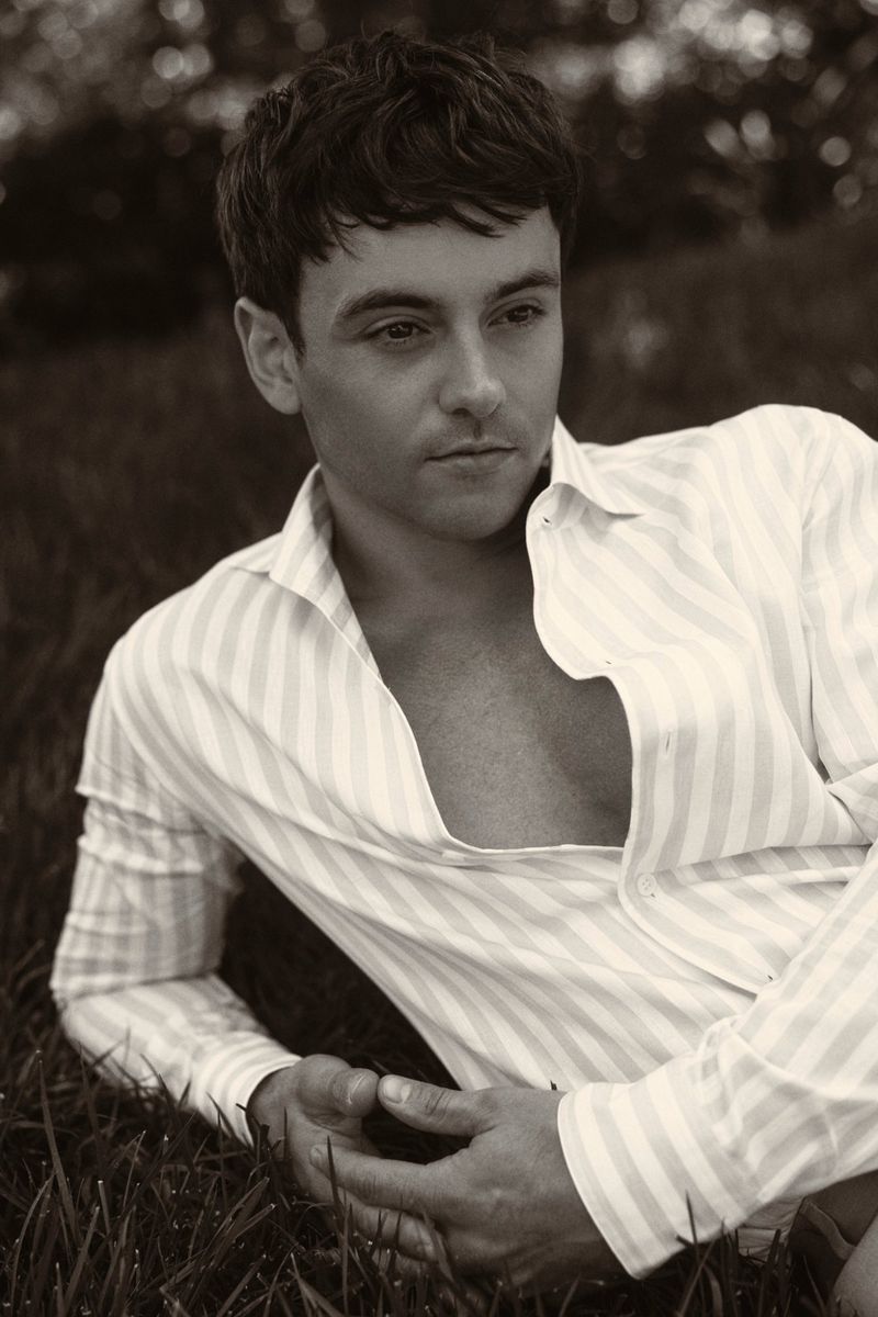 Portrait of a handsome adult male lying on green grass, wearing an unbuttoned striped shirt, photographed outdoors.