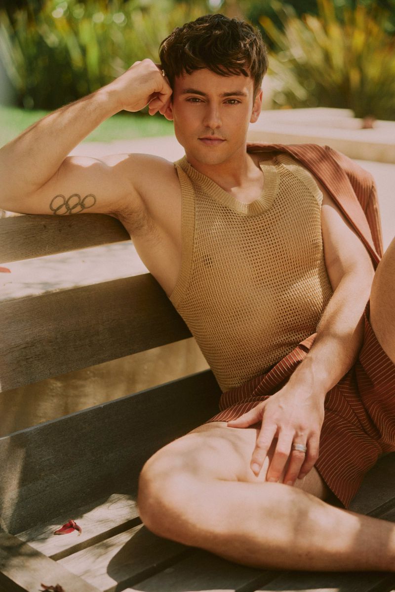 Portrait of adult male with Olympic rings tattoo, wearing knit tank top and jacket, sitting on wooden bench outdoors in natural light.