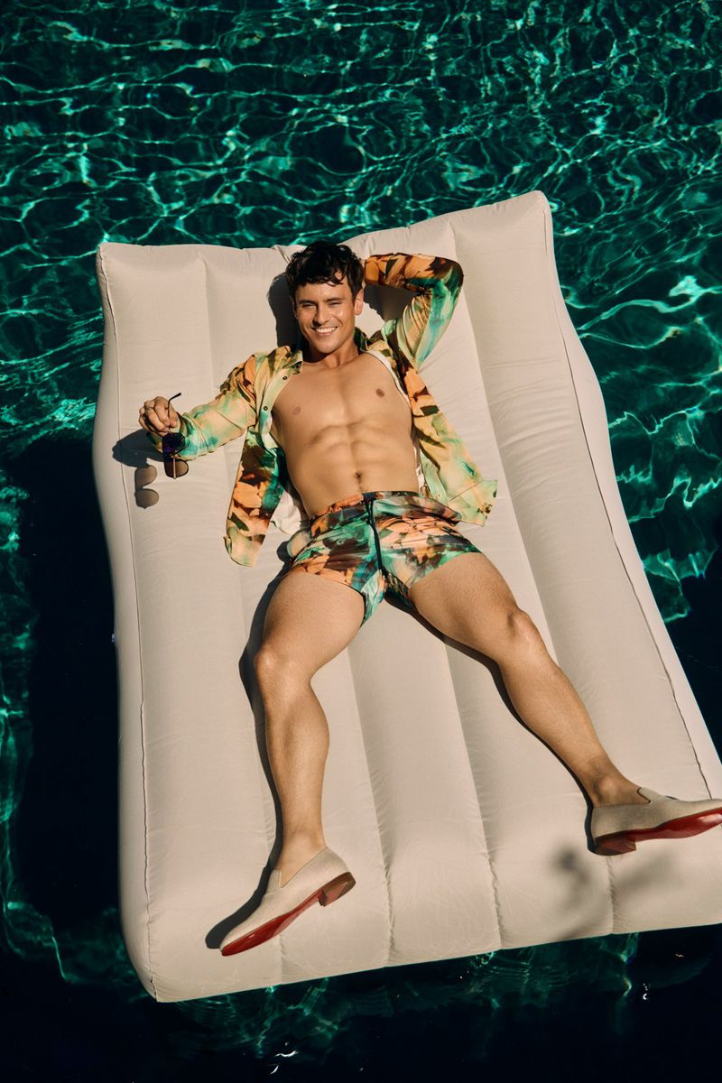 Man relaxing on a white inflatable raft in a swimming pool. He is wearing a floral shirt and swim trunks with loafers and sunglasses.