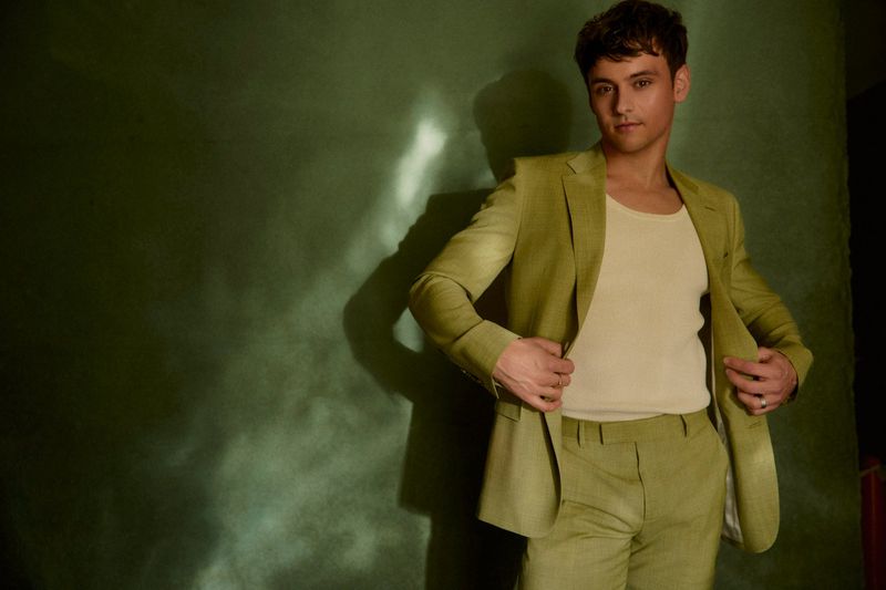 Portrait of a young adult man in a green suit and beige tank top standing indoors against a textured green wall in soft lighting.