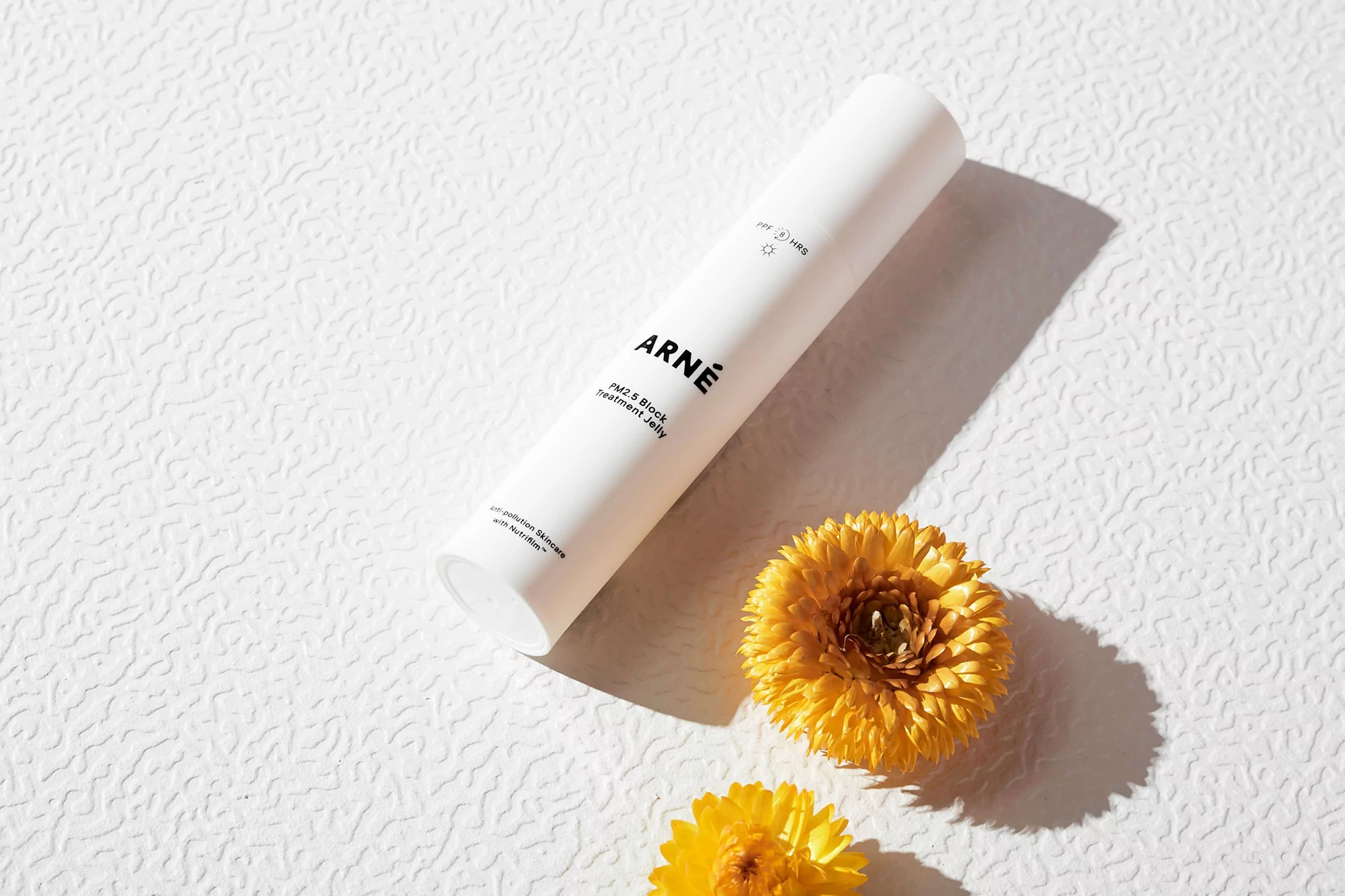 Close-up of white ARNE skincare bottle with yellow flowers, bright light, textured background for beauty and product advertising.