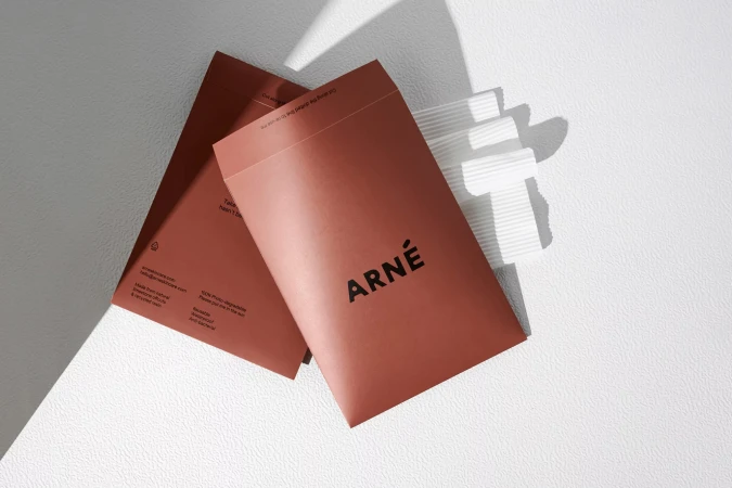 Two brown ARNE-branded envelopes with linear detail lay on a white surface in bright lighting, showcasing minimalist design and branding.
