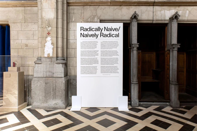 A white sign with the title "Radically Naive/Naively Radical" stands indoors, featuring columns and a black-and-white patterned floor.