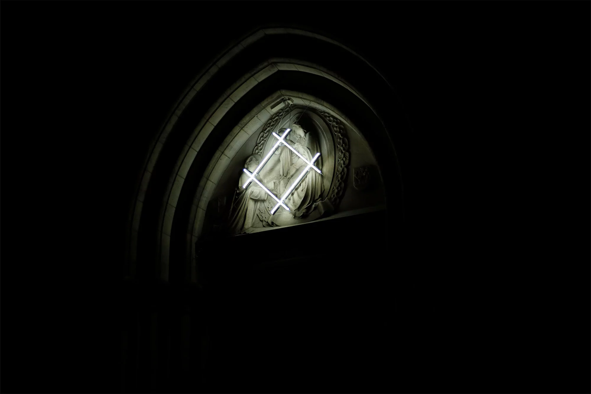 Sculptural art featuring a religious figure is set in a stone archway and is illuminated by a neon light installation.