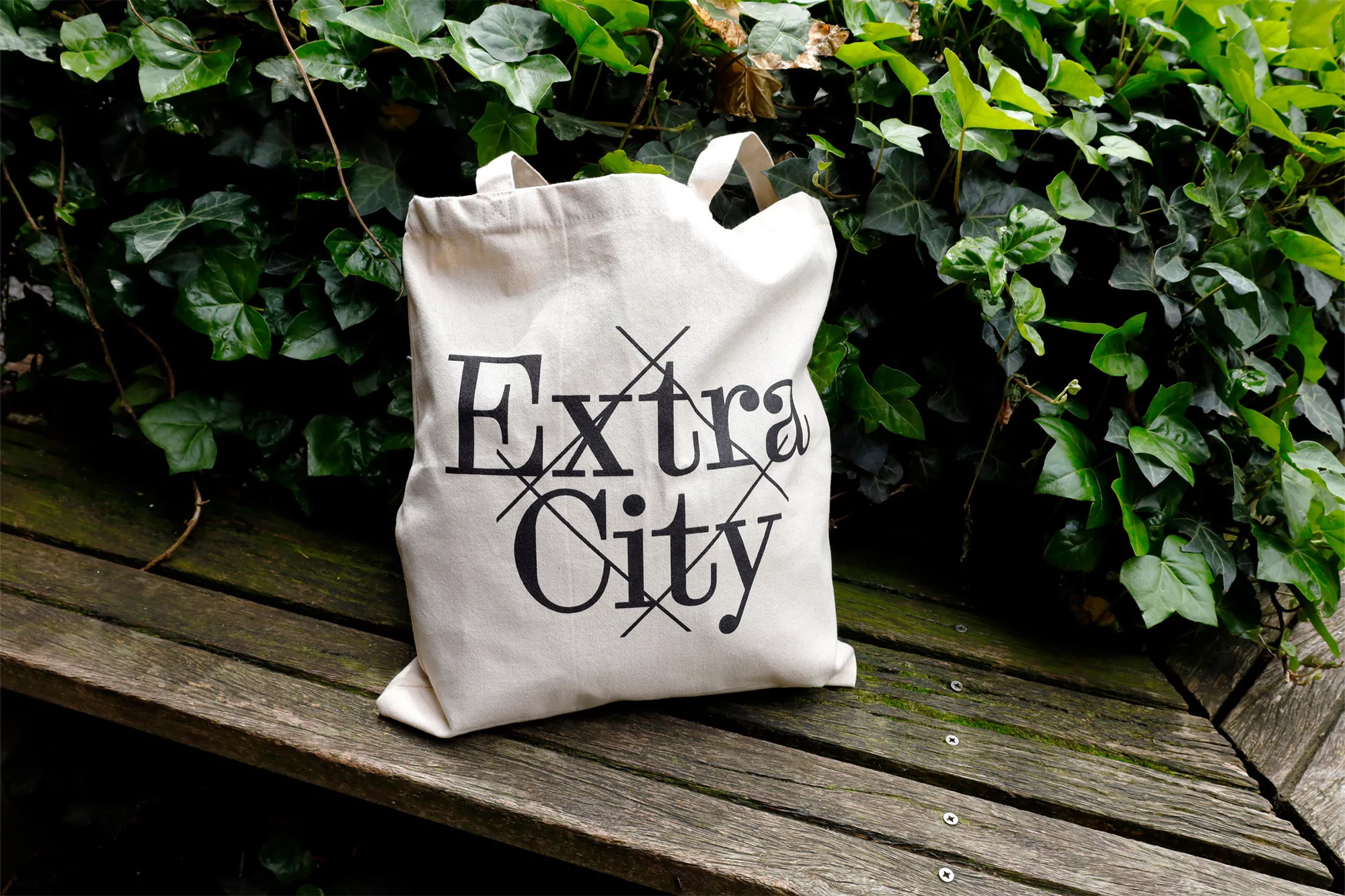 Canvas tote bag on a wooden bench, featuring 'Extra City' text, surrounded by vibrant green ivy, creating a natural outdoor setting.