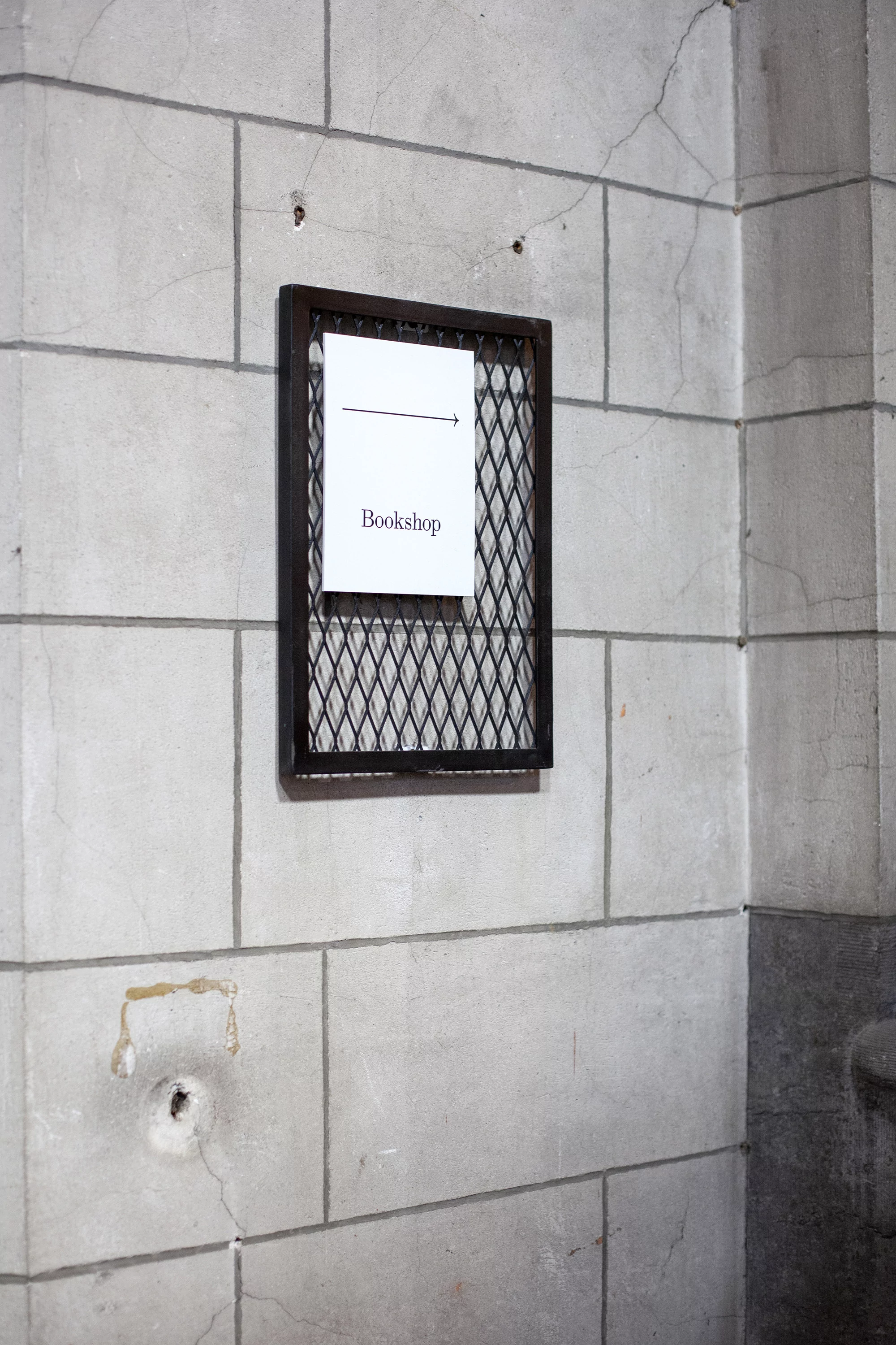 A sign with an arrow and the word 'Bookshop' hangs on a stone wall with visible cracks. The sign is enclosed in a metal frame.