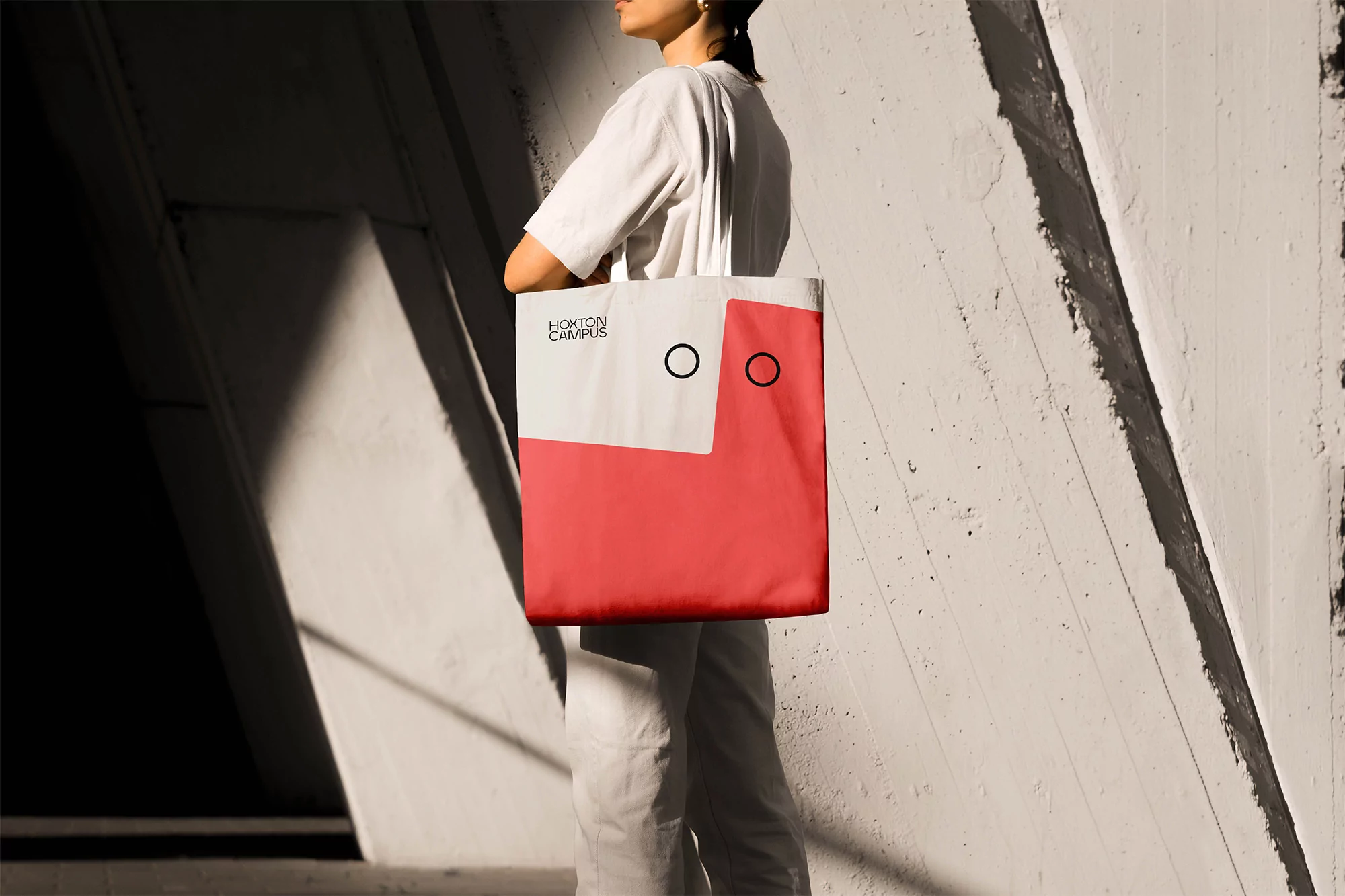 Woman carries a two-toned tote bag with circular designs against a concrete wall in sunlight, promoting campus or brand.