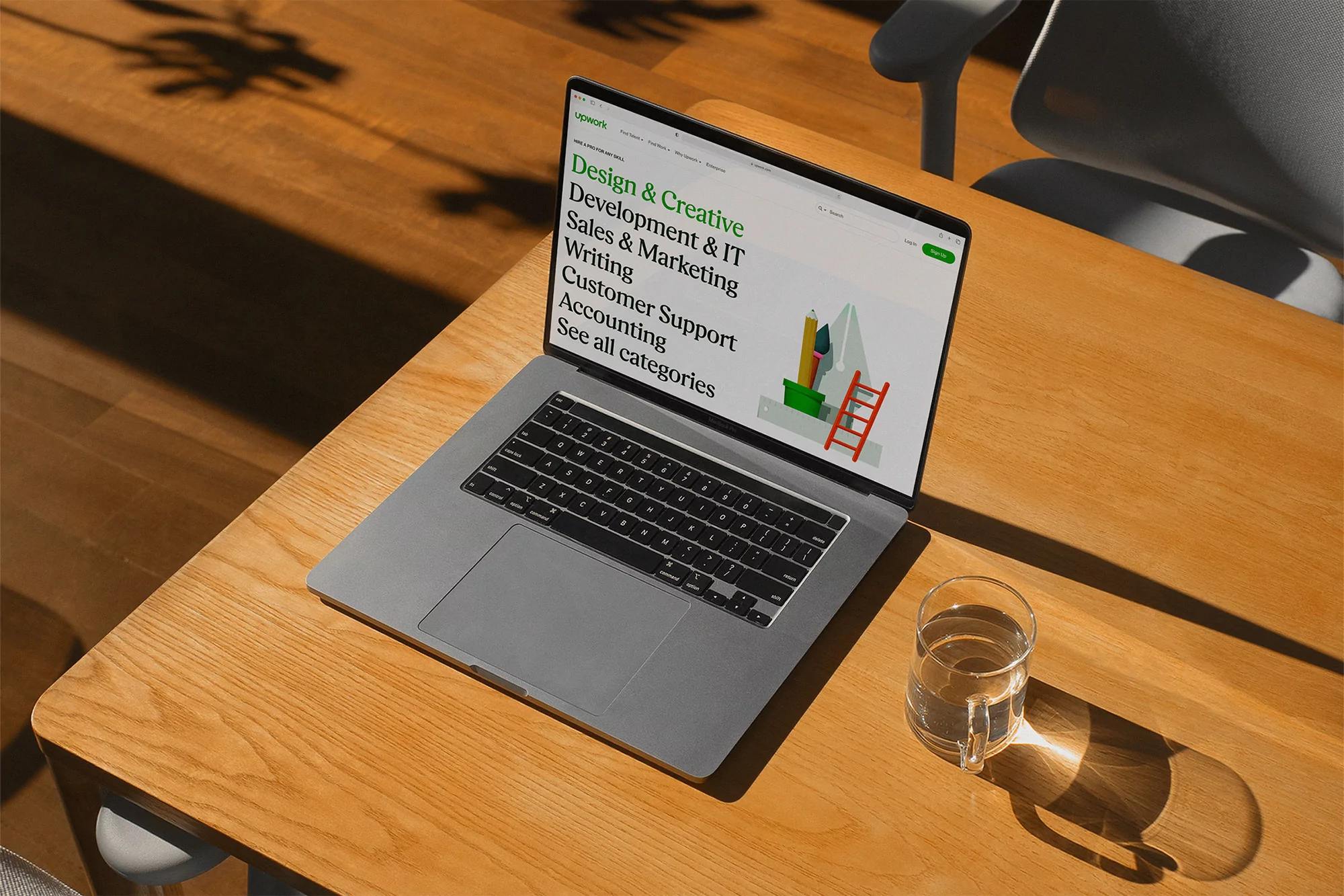 Silver laptop displaying a business website with a glass of water on a light wood table, illuminated by natural sunlight.