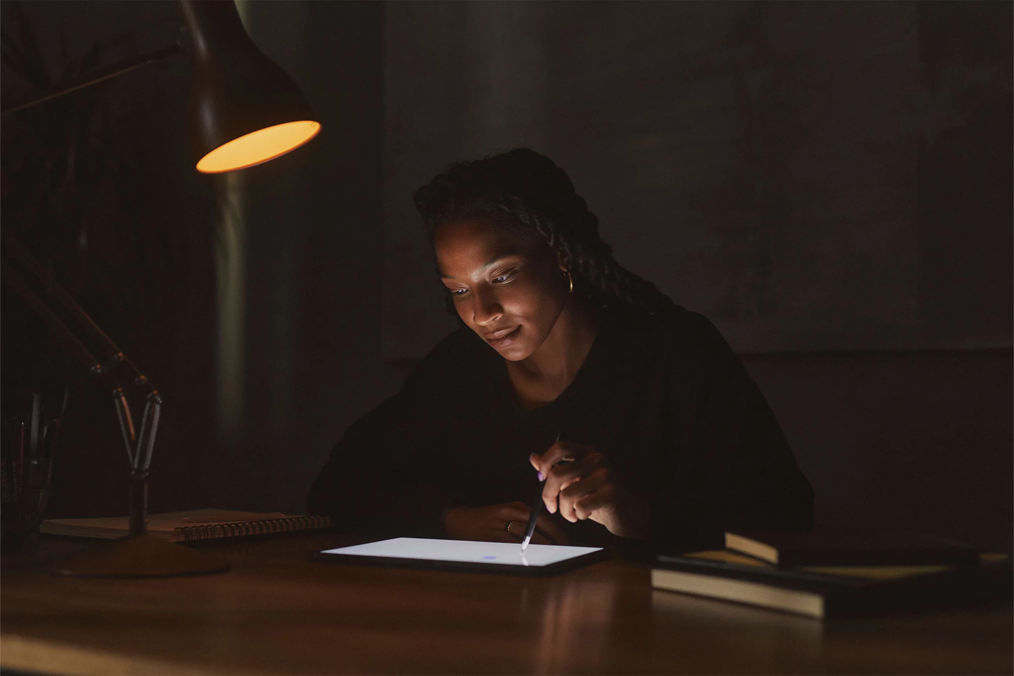 Woman with braids sits at desk in dim lighting, using stylus on tablet, illuminated by desk lamp; books and notebook nearby.