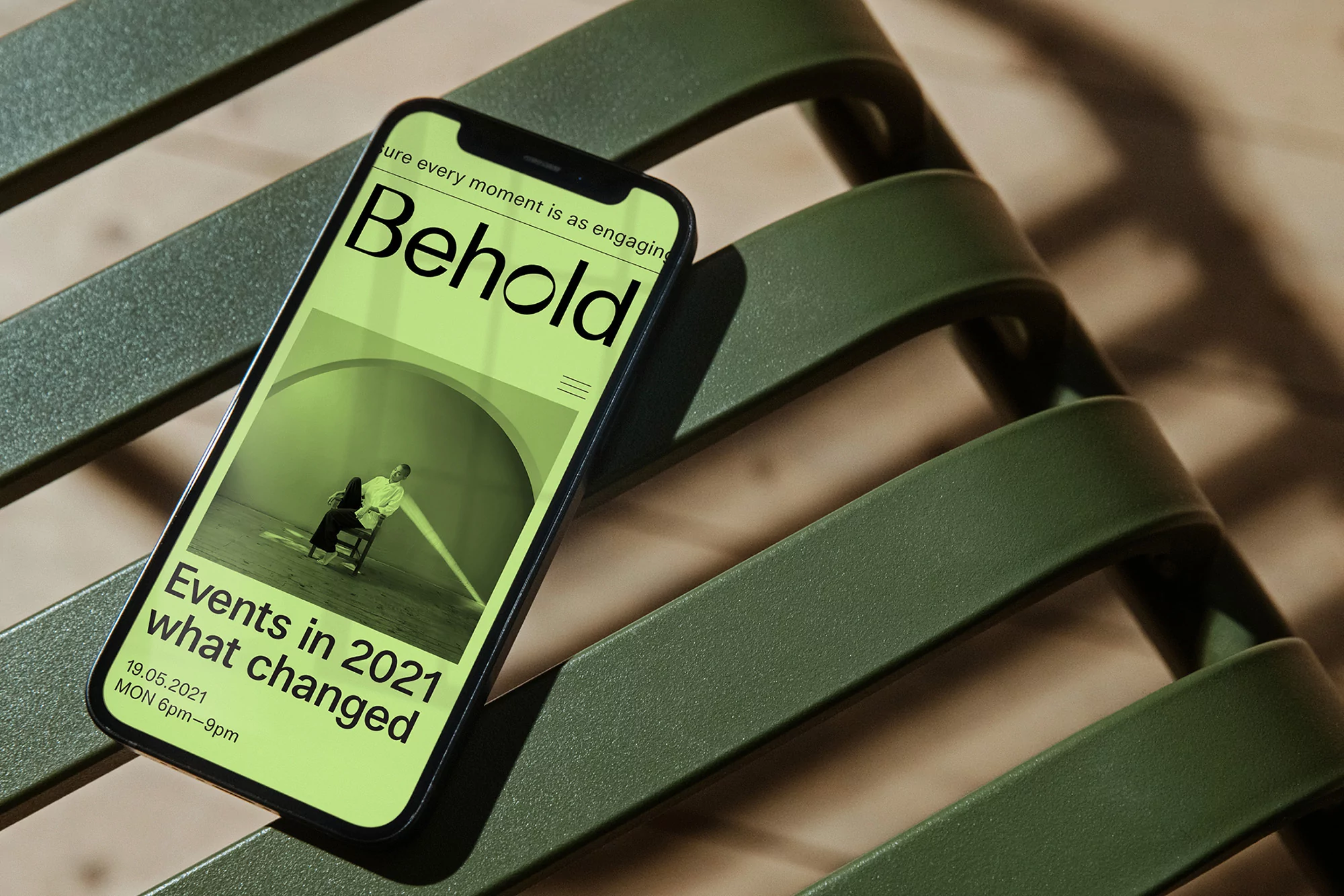 Smartphone displays a digital event poster on green slatted bench in close-up, promoting 'Behold' events in 2021.