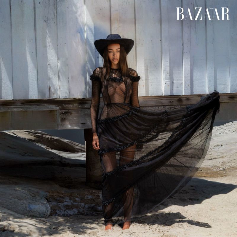 Fashion editorial shot of a woman in a black mesh dress and hat standing on a beach with a neutral color palette.