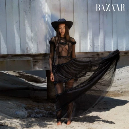 Fashion editorial shot of a woman in a black mesh dress and hat standing on a beach with a neutral color palette.