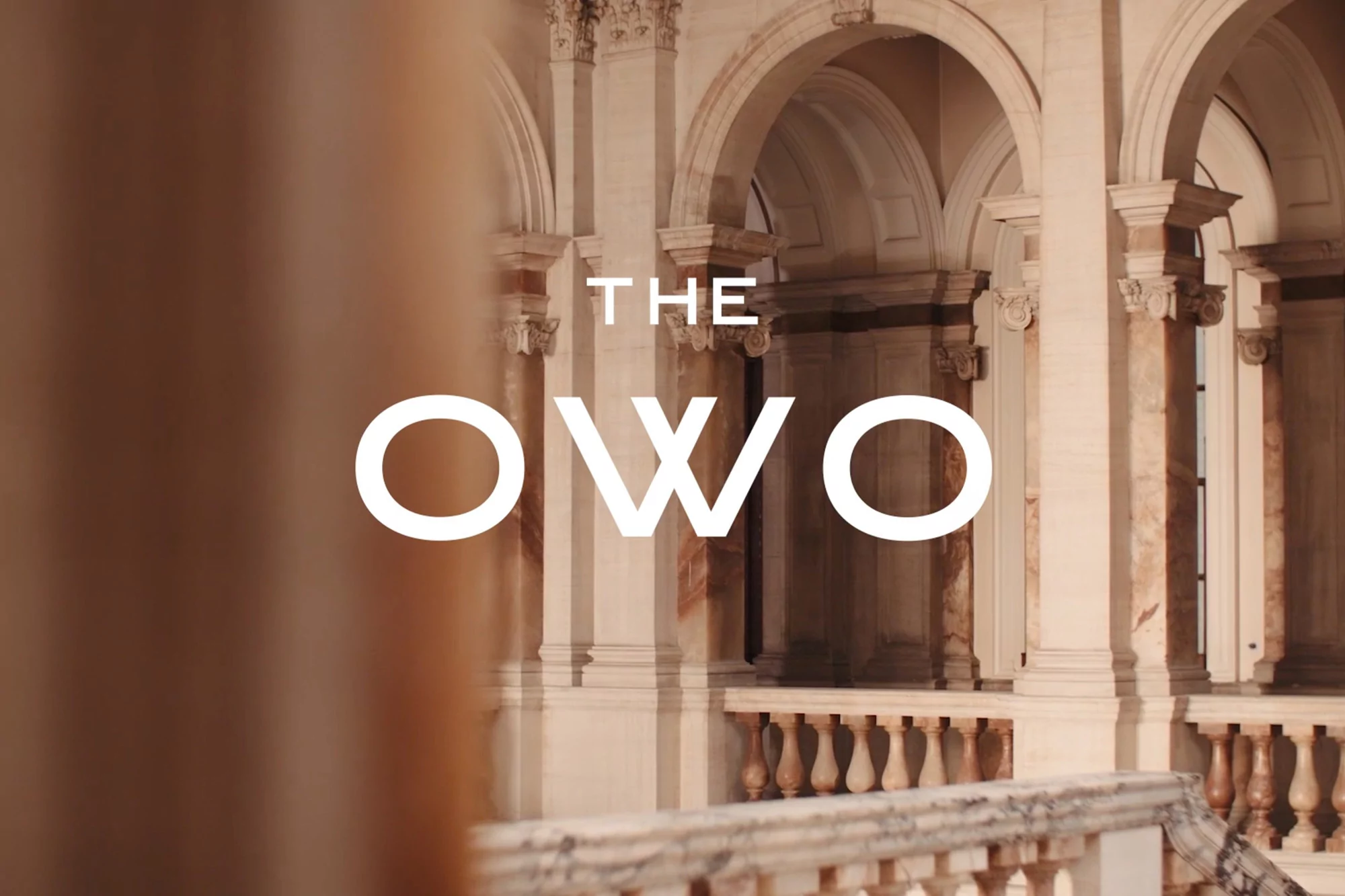 Architectural shot of an arched colonnade with ornate columns, stone balustrade, and the logo 'THE OWO' overlayed in white text.