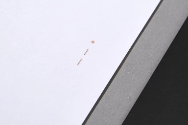 Close-up of debossed metallic copper logo mark on crisp white textured paper next to a grey edge.