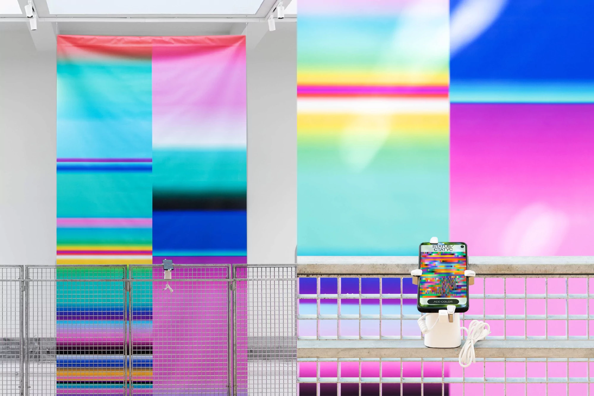 Abstract art exhibition with colorful graphic panels, metal barriers, and a smartphone on a railing displaying digital artwork.