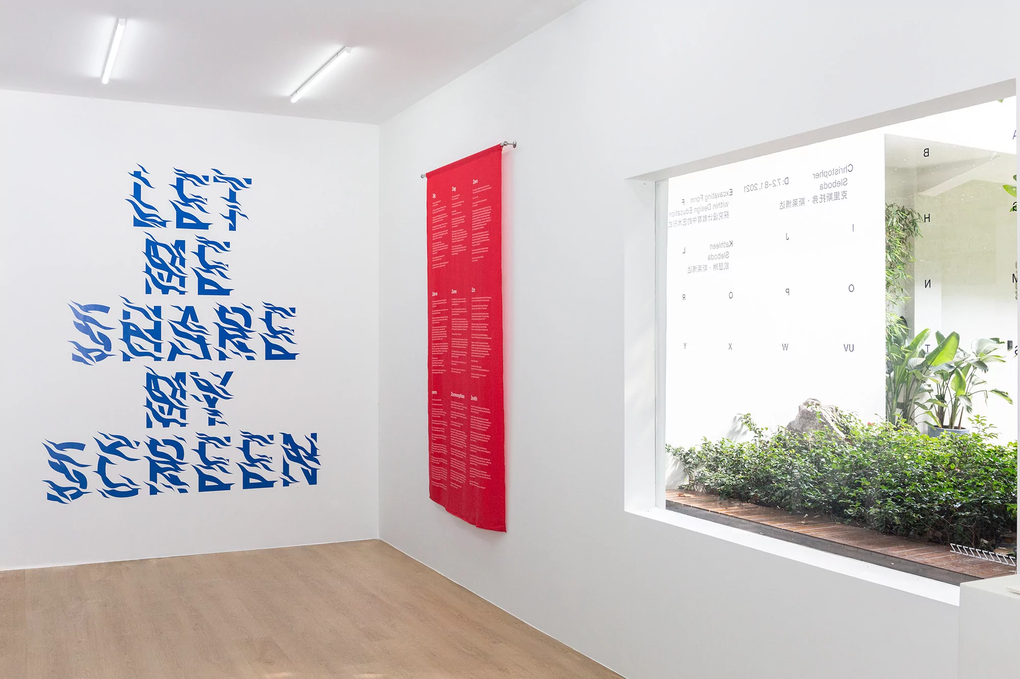 Minimalist art gallery features blue wall text, a red hanging banner, and a window view of a lush green outdoor garden.