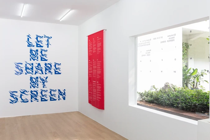 Minimalist art gallery features blue wall text, a red hanging banner, and a window view of a lush green outdoor garden.