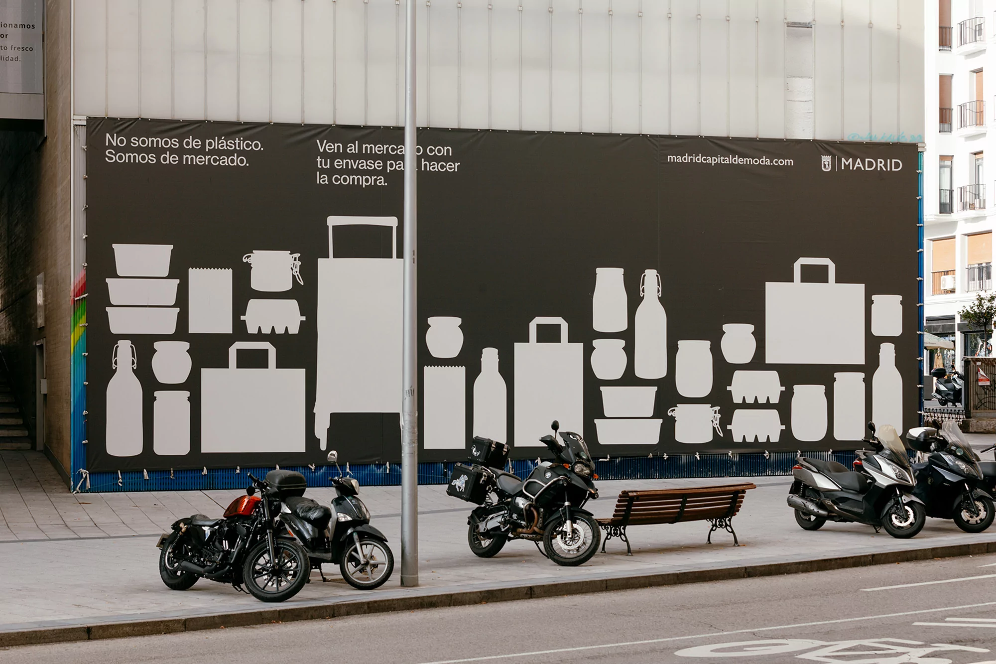 Motorcycles parked on a city street next to a large advertising banner featuring silhouettes of various shopping bags and containers.