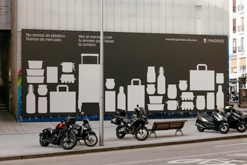 Motorcycles parked on a city street next to a large advertising banner featuring silhouettes of various shopping bags and containers.
