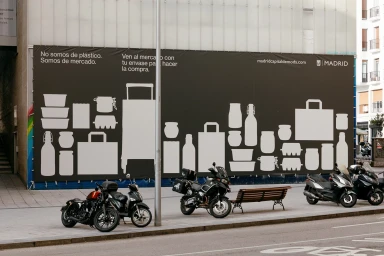 Large dark billboard on a city street displaying white graphic icons of containers and shopping bags with Spanish text.