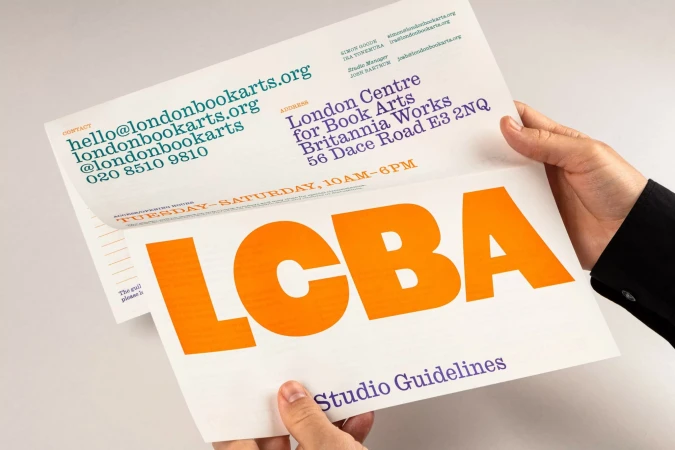 Two hands hold a London Book Arts flyer with contact info, address, and hours. The flyer displays the orange letters LCBA and 'Studio Guidelines'.
