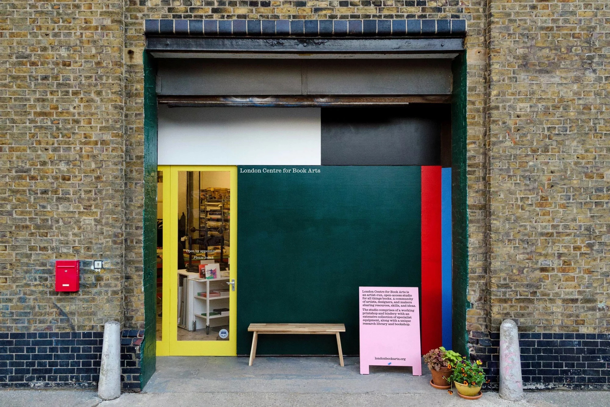 Exterior of London Centre for Book Arts shows the entrance, a bench, potted plants, and a pink sign in front of a brick building.