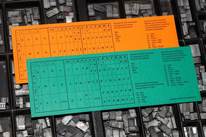 Orange and green cards detailing typeface spacing sit in a letterpress tray filled with metal type blocks, high-angle shot, detailed.