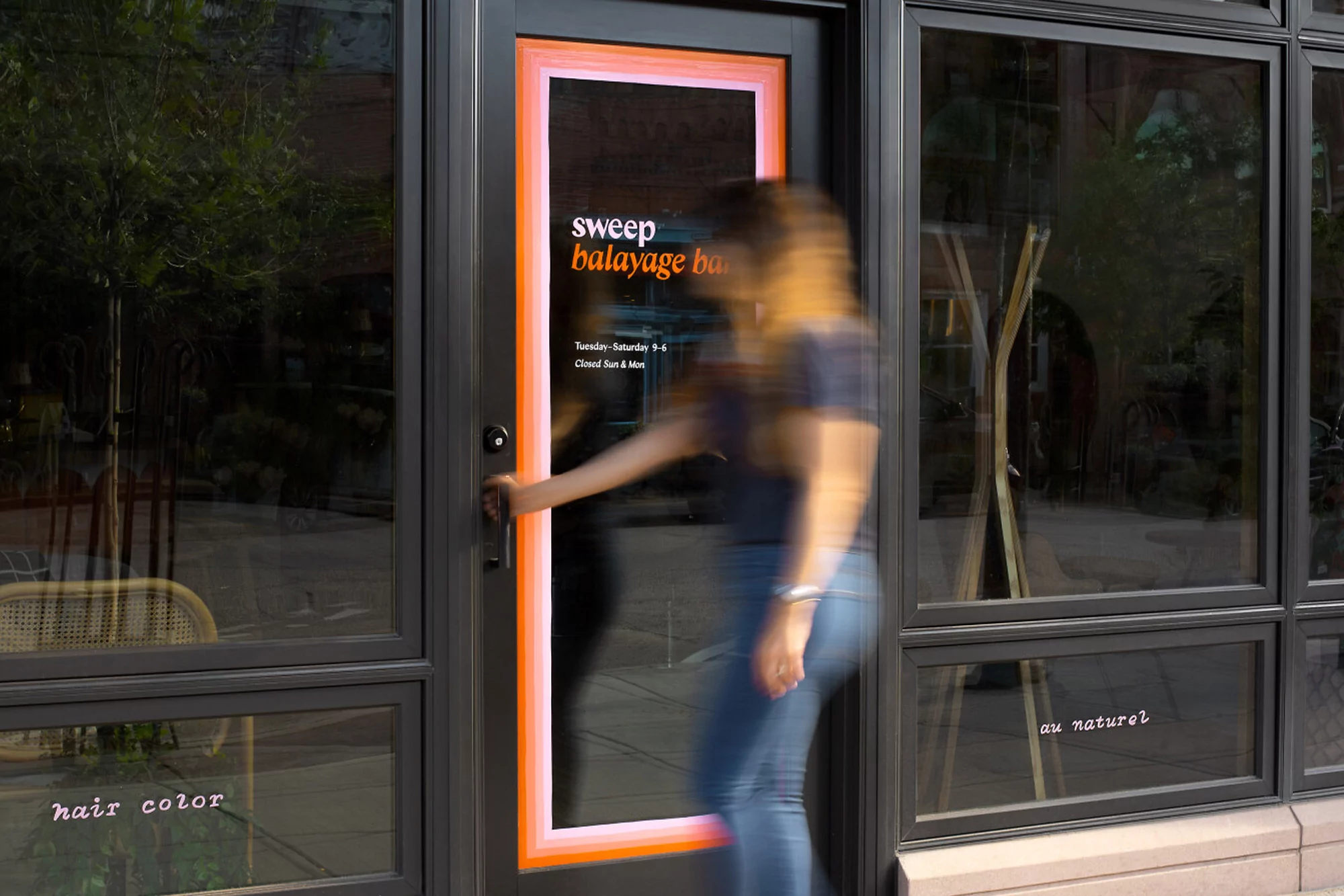 Woman enters a hair salon with black framed windows and doors, featuring salon signage and a modern, urban setting, with neutral colors.