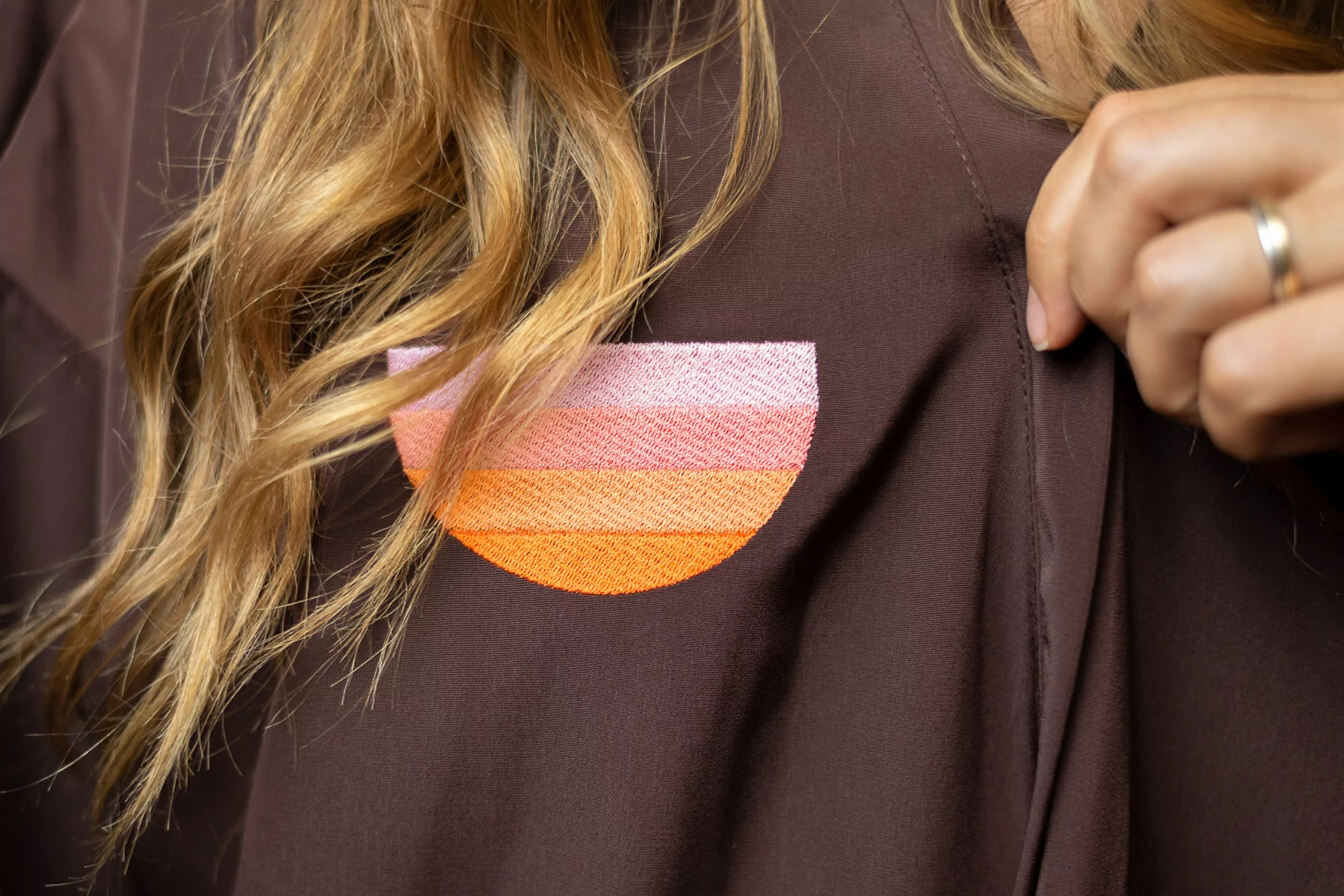 Closeup of a person wearing a brown top with an embroidered, horizontal striped design, revealing blonde hair and hand.