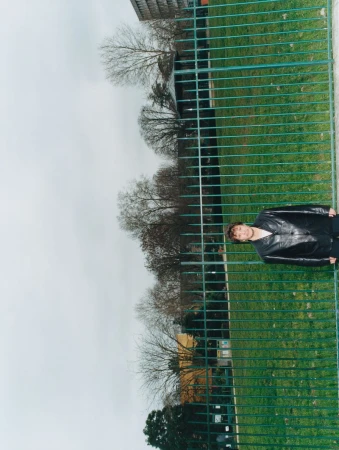 Person standing behind a green metal fence in a park on an overcast day with bare trees and an apartment building in the background.