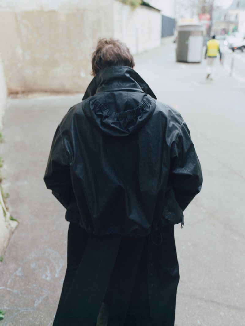 Back view of a person walking on a city sidewalk wearing a black hooded coat and pants, with blurred buildings and a street in the background.