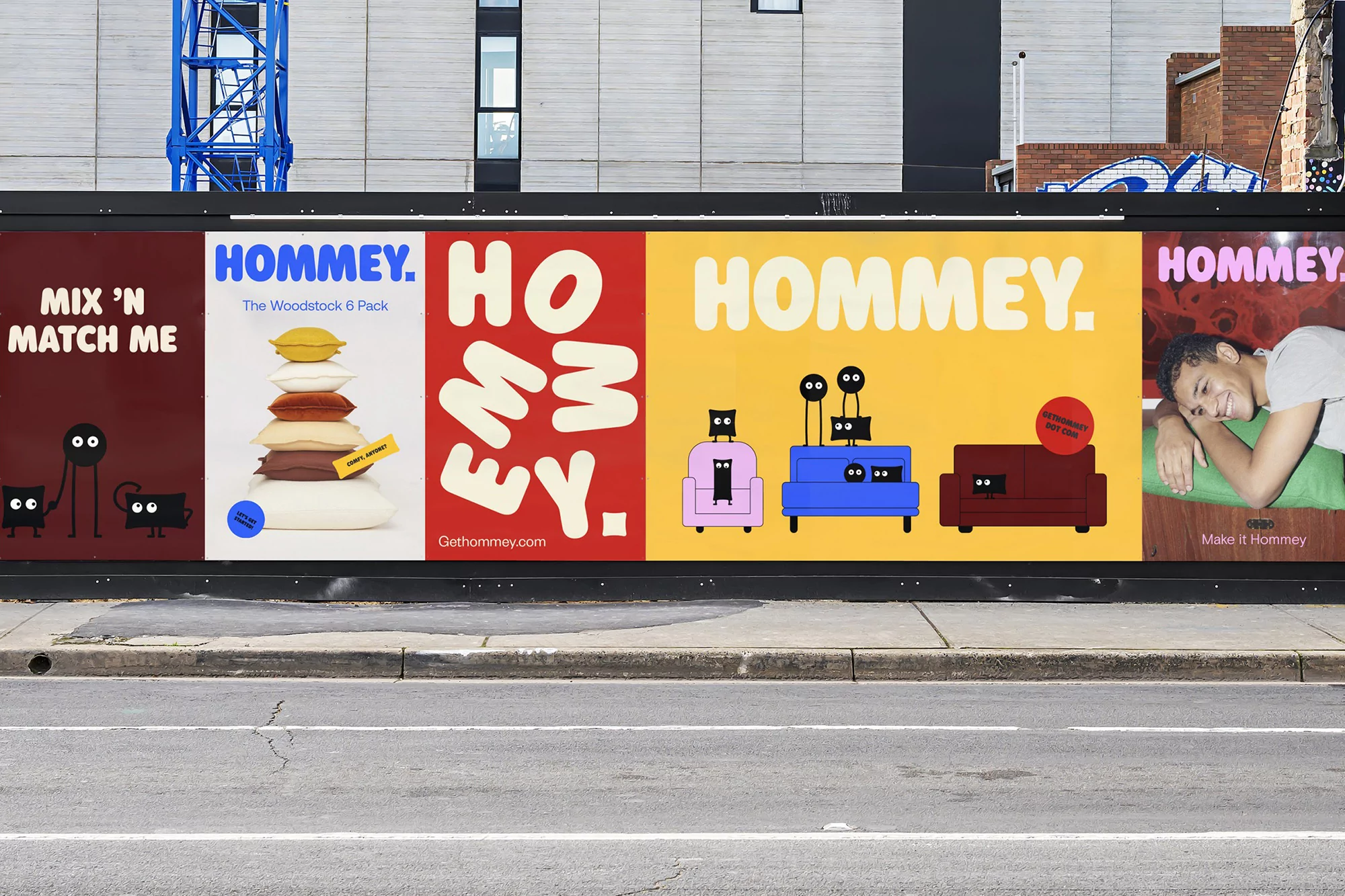 Eye-level view of Hommey advertising billboard featuring pillows, furniture, abstract characters, and a smiling person, on an urban street.