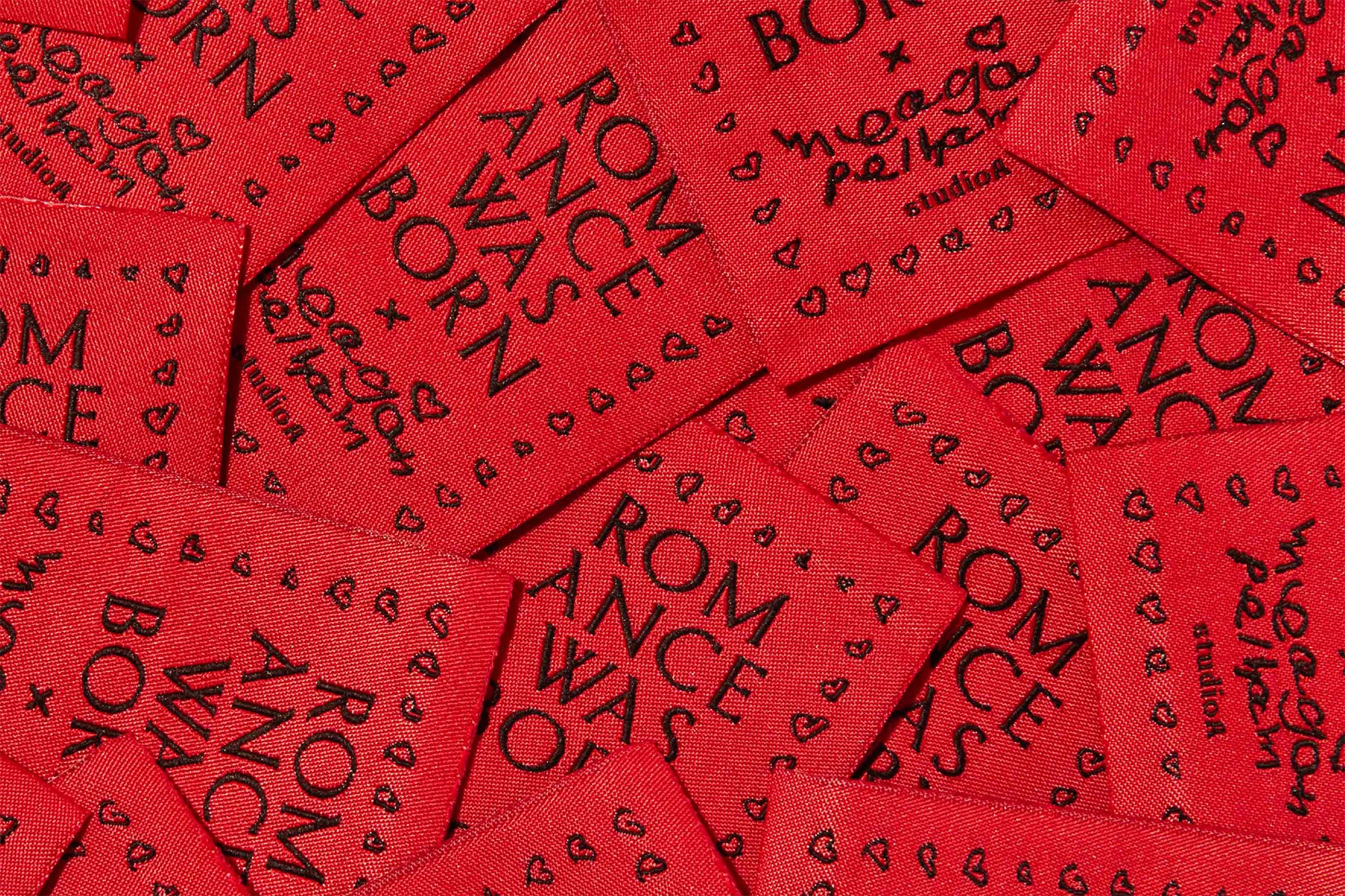 Macro shot of red fabric labels with black text that reads 'Romance Was Born' and 'meagan pelham studios' repeated across surface.