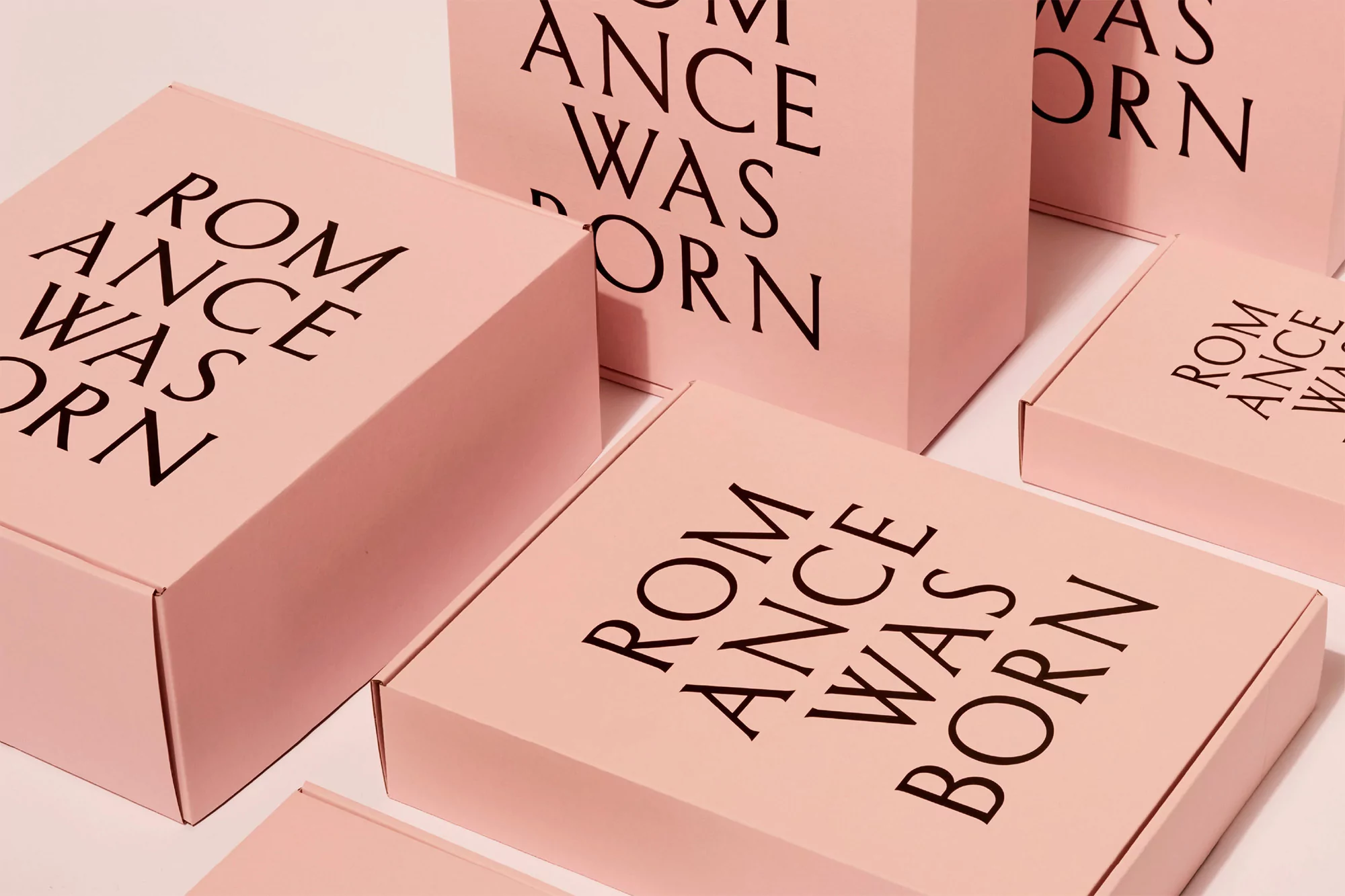 Stacks of pastel pink boxes and bags with bold black text 'Romance Was Born' create a minimalist, fashion-forward product presentation.