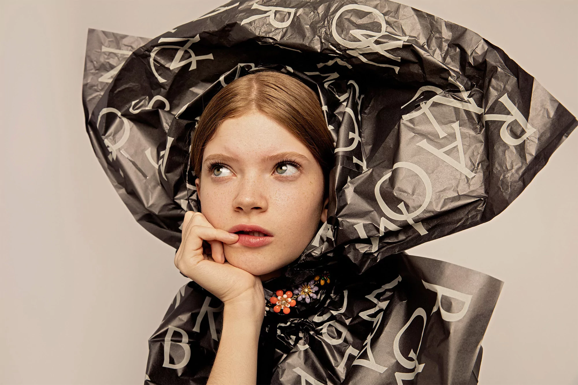 Young adult female portrait with light skin and red hair, posing in a headpiece made of black paper with white lettering.