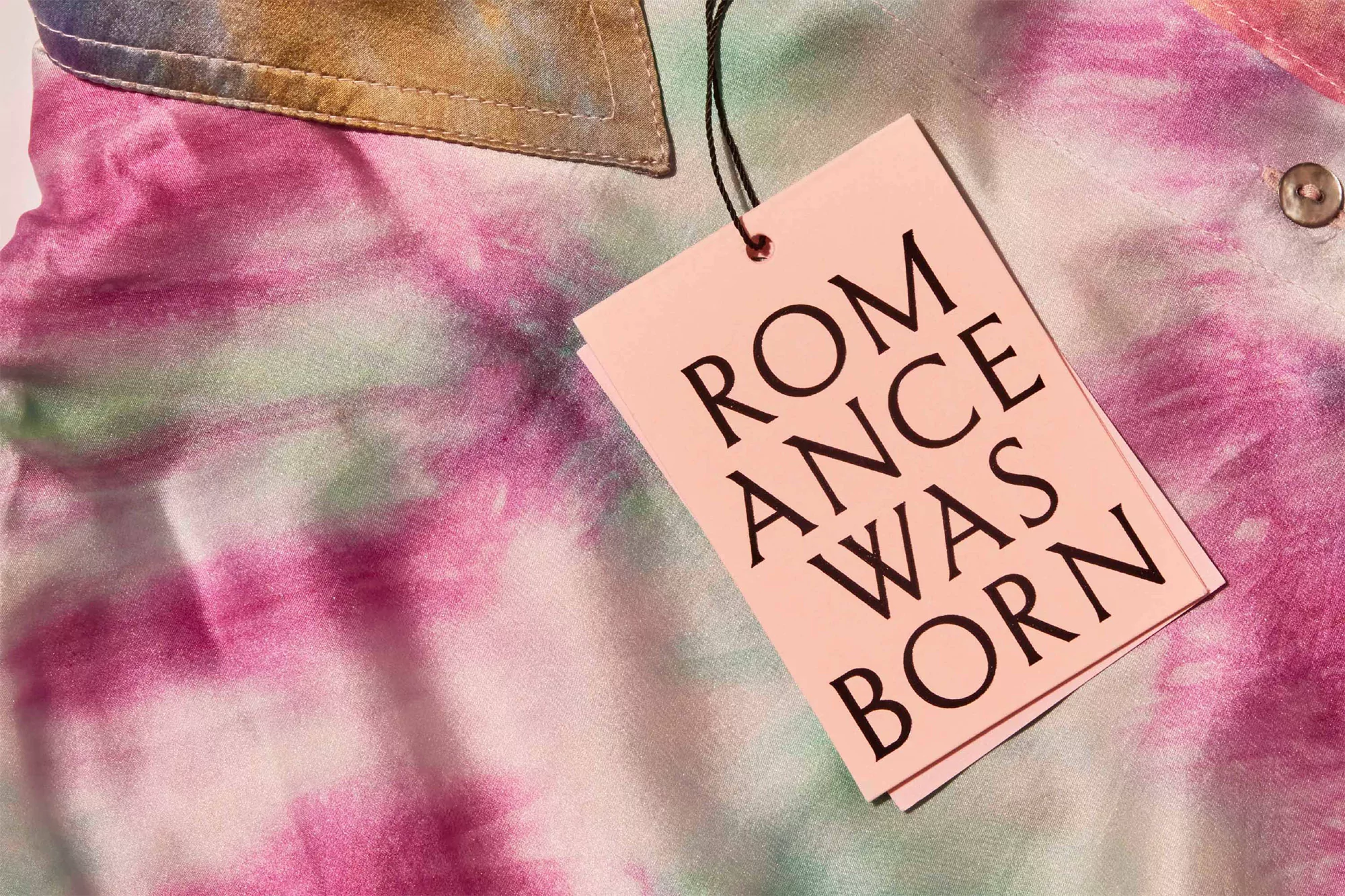 Close-up of a colorful tie-dye shirt featuring hues of pink, white, and green, paired with a pale pink tag with black text.