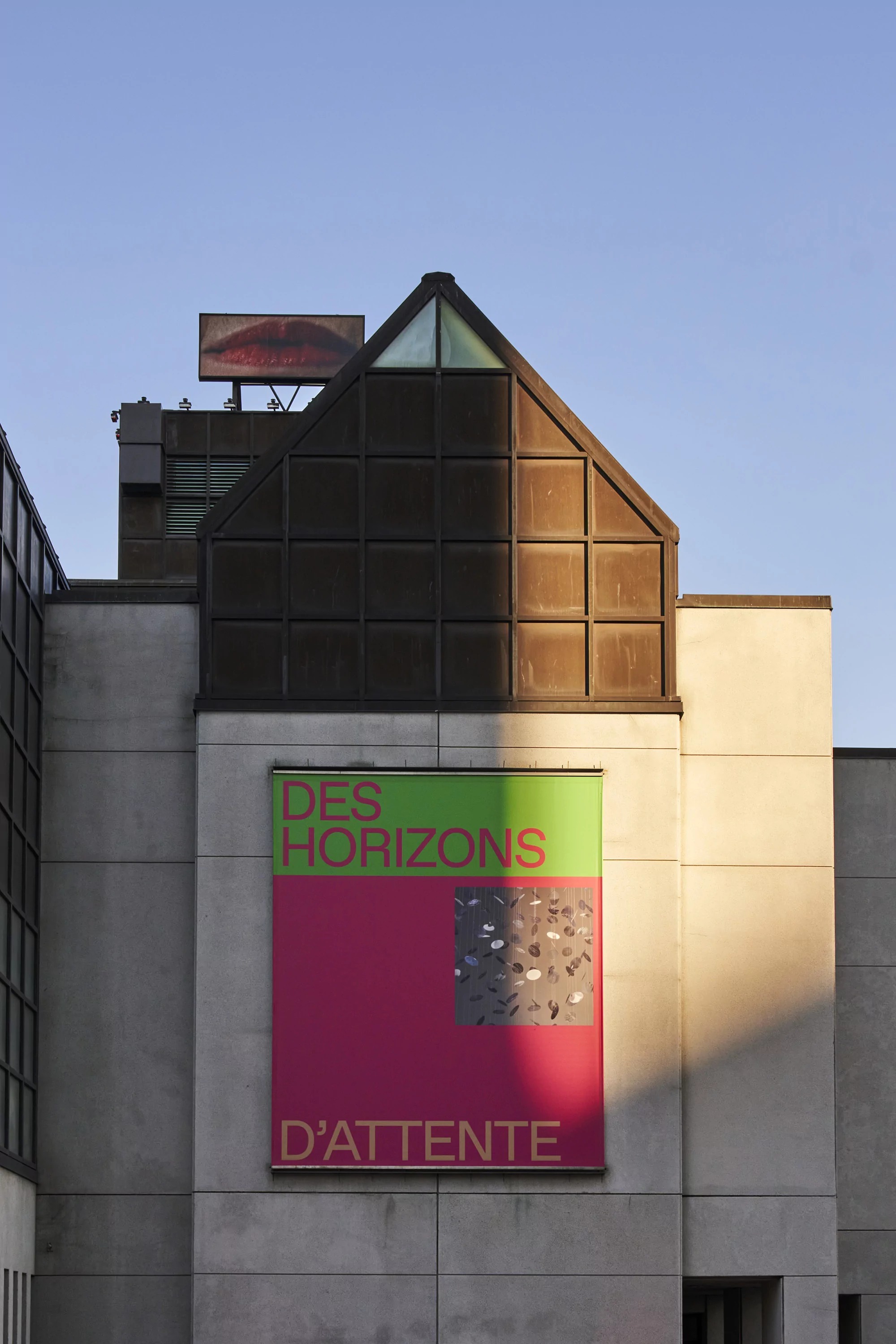 Modern building facade with an art exhibition poster featuring bold text and graphic elements under a clear blue sky.
