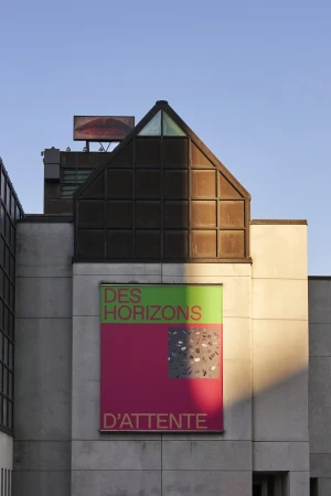Modern building facade with an art exhibition poster featuring bold text and graphic elements under a clear blue sky.